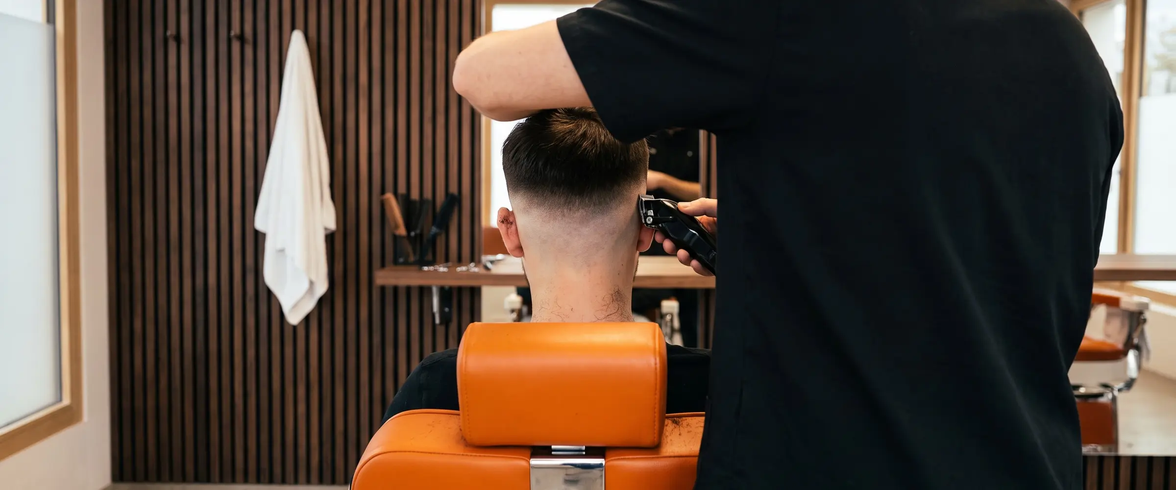 Barber in schwarzem Kasack setzt am Hinterkopf eines Kunden einen Skin Fade, orangefarbener Stuhl, dunkle Holzlatten