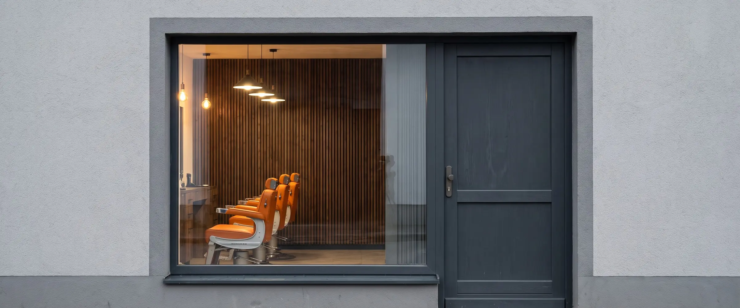 Ladenfront mit großem Fenster und grauer Tür, drinnen drei orangefarbene Stühle vor Holzlatten